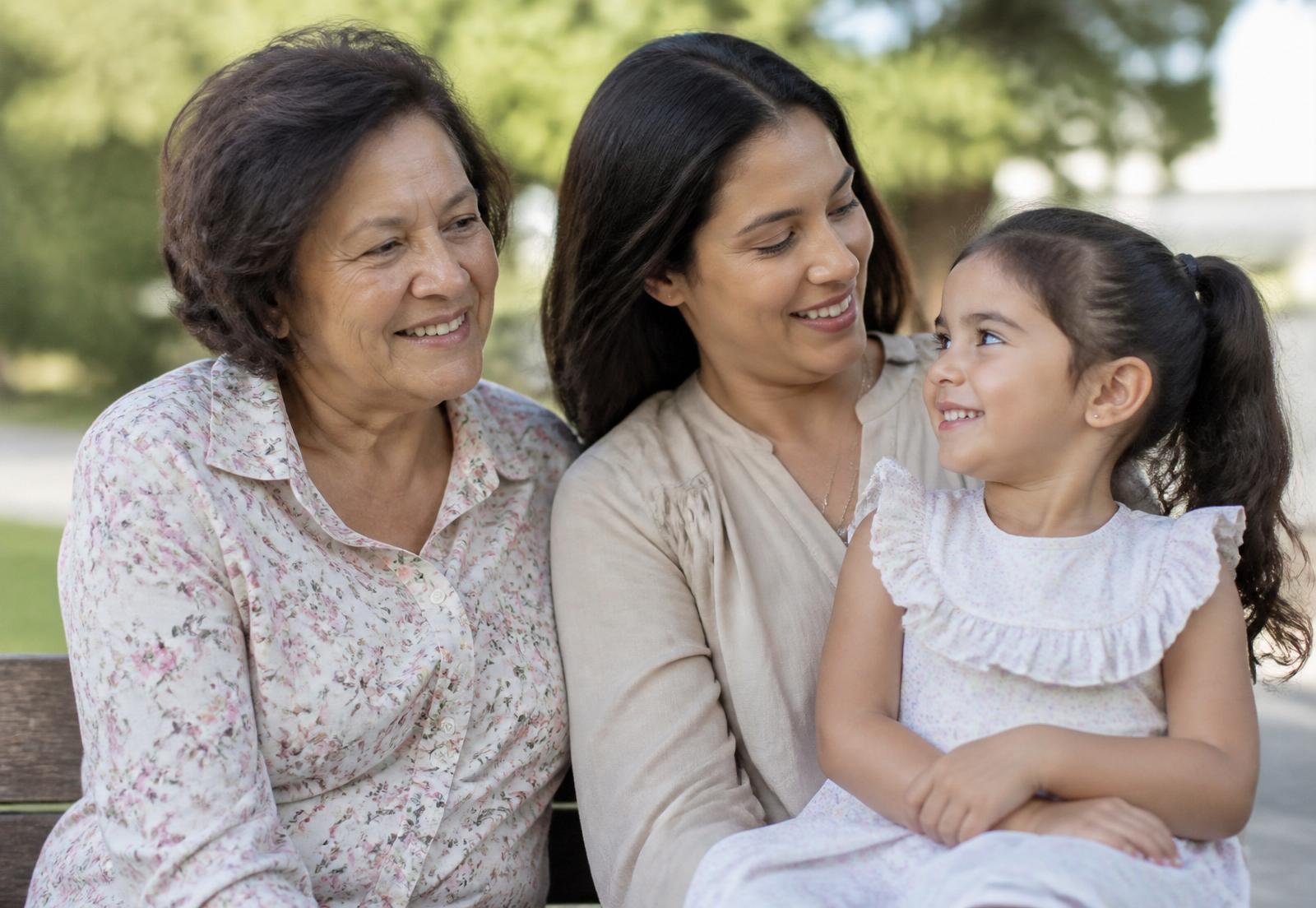 A grandmother, mother, and young daughter laughing together on a sunlit porch.
